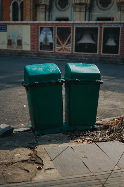 A large blue plastic wheelie bin with a textured surface and a slightly worn appearance is positioned on the pavement beside a white delivery van or mobile unit, which features a sliding door, visible