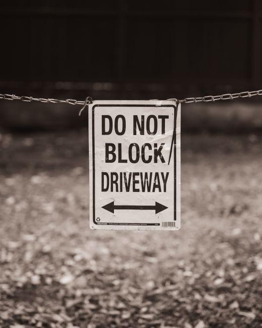 A wooden pallet with a weathered and slightly warped surface leans against a white brick wall on a sidewalk, positioned below a red and white 'No Parking in This Area' sign mounted directly above it.
