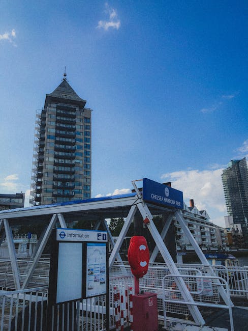 The image depicts a section of Chelsea Harbour Pier with a clear blue sky overhead and a few scattered clouds. In the foreground, there is a white metal structure supporting the pier entrance, topped