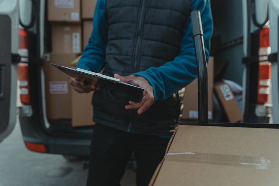 A person wearing a blue beanie and red jacket is standing outside a modern, white commercial building with large glass windows. They are stacking several cardboard boxes next to a small, silver delive